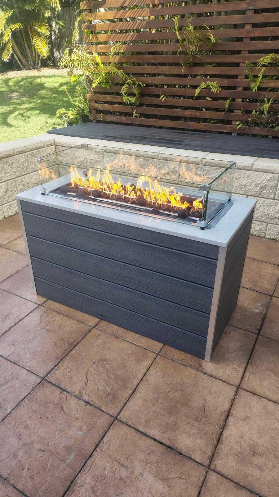 Custom-made gas fire pit with charcoal Modwood cladding and natural stone top burning in a Brisbane back garden